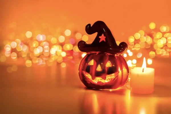 Zucca di Halloween luminosa con cappello da strega e candela accesa su sfondo sfocato arancione.